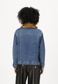 Denim jacket featuring a brown faux fur collar, relaxed fit, and classic stitching details, viewed from the back. Black leather pants visible.