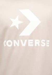 Ανοιχτό μπεζ βαμβακερό μπλουζάκι με μεγάλο λευκό αστέρι, σχέδιο βέλους και έντονη λευ κή επιγραφή "CONVERSE" στο κάτω μέρος.