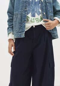 Giacca di denim con lavaggio azzurro chiaro, felpa bianca con grafica blu e verde, e pantaloni cargo blu scuro con tasche laterali e pieghe.