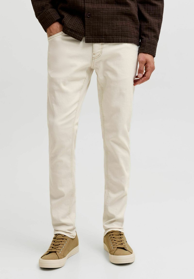 Homme portant un pantalon slim beige clair, une chemise à carreaux marron et des baskets basses couleur fauve, debout, une main dans la poche.