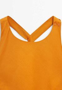Vêtement orange sans manches avec des bretelles croisées attachées par deux boutons sur une épaule, sur fond blanc.