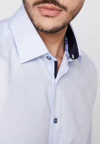 Homme portant une chemise à carreaux bleu clair avec doublure intérieure de col bleu foncé et boutons bleus, montrant partiellement le visage et le cou.