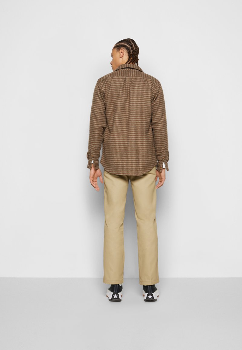 Chemise à carreaux marron à boutons avec des manches longues, accompagnée de pantalons beige clair et de baskets noires avec des étoiles blanches.