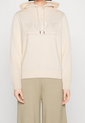 Cremefarbener Kapuzenpullover mit Kordelzug und geprägt "GANT American Sportswear"-Logo, getragen zu beigen Hosen mit weitem Bein.