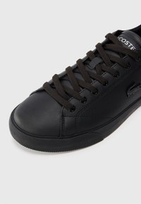 Sapatilha de couro preto com biqueira arredondada, atacadores castanhos e sola de borracha texturizada preta. O logo da Lacoste está exibido na lateral.