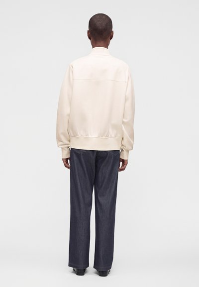 Personne portant une veste crème et un pantalon large bleu foncé, debout de dos face au spectateur, sur un fond clair uni.