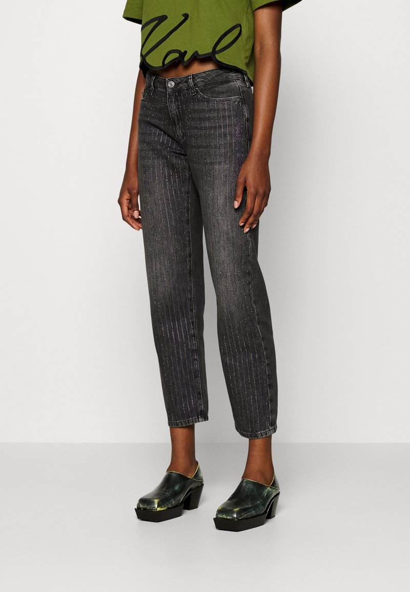 Black denim jeans with vertical metallic pinstripes, tapered leg, high waist, and a cropped green top with bold black lettering. Black clogs with green accents.