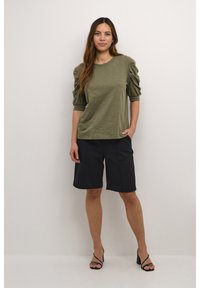 Olīvu zaļš puff-sleeve tops ar apaļu kakla izgriezumu, savienots ar ceļgala garuma melnām šortiem un melniem siksniņu augstpēdu sandales.