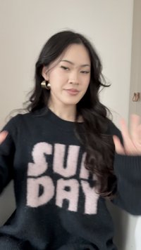 Ženska z dolgimi temnimi lasmi, ki nosi zlate obročaste uhane in črni pulover z napisom "NEDÉLJA" v velikih roza črkah, dviguje roke pred bež zidom.