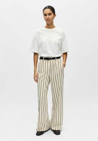 T-shirt blanc oversize, pantalon large beige à rayures verticales et espacées uniformément, ceinture noire, et chaussures noires.