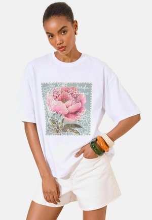 Nő fehér, túlméretezett pólóban, pink virággal és a „Jardin Botanique” felirattal, fehér rövidnadrágban, színes karkötőkkel az egyik csuklóján.