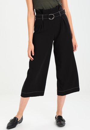 Mujer con pantalones negros de pierna ancha y cortos con cinturón y mocasines negros de punta afilada sobre un fondo blanco.