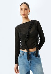Schwarzes Oberteil mit langen Ärmeln, das an einer Seite Rüschendetails aufweist und aus transparenter Materie gefertigt ist. Kombiniert mit blauen Jeans in einer cropped Ausführung.