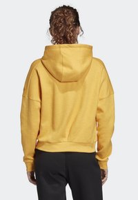 Žlutá mikina s kapucí v uvolněném střihu, stahovací šňůrkou na kapuci, žebrovanými manžetami a lemem. Má měkkou strukturu a spadlé švy na ramenou.