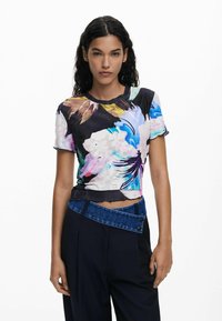 Mujer con cabello rizado negro que lleva una blusa corta de manga corta de flores multicolores y pantalones negros de cintura alta con una banda de mezclilla en ángulo.