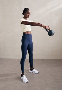 Set za aktivno odijelo: svijetložuta kratka majica s perforacijom, mornarsko plave tajice viskog struka i bijele tenisice s mornarskim detaljima, drži kettlebell.