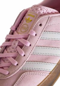Pink suede sneaker with white leather stripes, flat laces, textured tongue, gold branding, and a clear rubber sole.