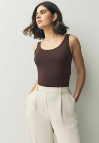 Ruskea ilmanhiha tank top, joka on yhdistetty korkeavyötäröisiin, vaaleanbeigeihin housuihin. Sileä kangas, jossa on istuva siluetti ja hienovarainen tekstuuri.
