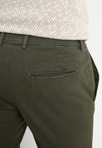 Primo piano di pantaloni verde scuro con una tasca posteriore a filetto, indossati con un maglione in maglia testurizzata beige chiaro.