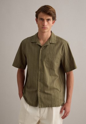 Jonge man met bruin haar die een olijfgroen overhemd met korte mouwen en knopen en crèmekleurige broek draagt tegen een effen achtergrond.