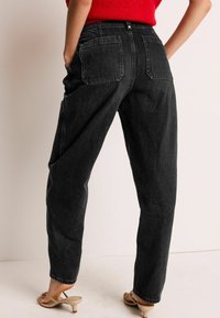 Pantaloni din denim negri, cu talie înaltă și croi lejer, având două buzunare la spate și cusături minime. Purtați cu sandale bej.