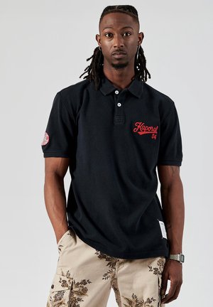 Zwarte polo shirt van katoen, met een rood geborduurd logo en een knoopsluiting met twee witte knopen; gecombineerd met beige gestreepte shorts.