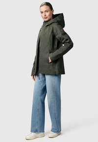 Femme portant une veste à capuche vert foncé, un jean large bleu clair et des baskets blanches, debout avec une main dans la poche de la veste.