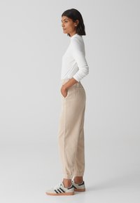 Beige corduroy joggers met elastische boorden en zakken, gecombineerd met een witte langemouwen top en grijze sneakers met zwarte strepen.