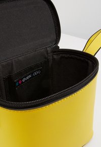 Trousse de maquillage en cuir jaune avec une fermeture éclair noire, intérieure en tissu noir et une étiquette de la marque Even & Odd.