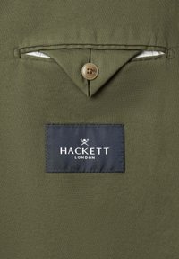 Olivgrüner, strukturierter Stoff mit einer geknöpften Klappentasche über einem auf dem Material aufgenähten schwarzen Hackett London-Label.