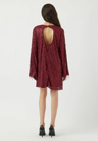 Viininpunainen mekko, jossa pitkät hihat, takaosassa avonainen keyhole-leikkaus ja hapsureunus. Sileä pinta kiiltävällä viimeistelyllä. Mustat korkokengät.