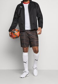 Sort vindjakke, hvid skjorte, brune stribede shorts, hvide sokker og hvide fodboldstøvler, der holder en orange og sort fodbold.