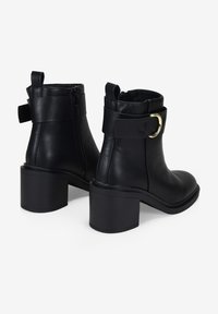 Botas de cuero negro hasta el tobillo con punta redonda, tacón cuadrado y cremallera lateral. Presentan una correa con un detalle de hebilla en tono dorado. Textura suave.