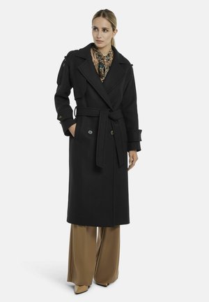 Frau trägt einen schwarzen, langen Mantel mit Gürtel, braune weite Hosen, beige Schuhe und ein gemustertes Halstuch, steht mit einer Hand in der Tasche.