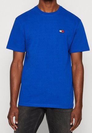 Sininen puuvillainen T-paita, jossa on pyöreä kaula-aukko, lyhyet hihat ja pieni brändilogo-painatus punaisella, valkoisella ja mustalla rinnassa.