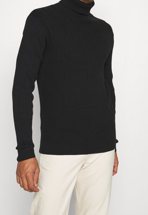 Mężczyzna ubrany w czarny prążkowany golf i kremowe spodnie, stojący na jednolitym jasnym tle.