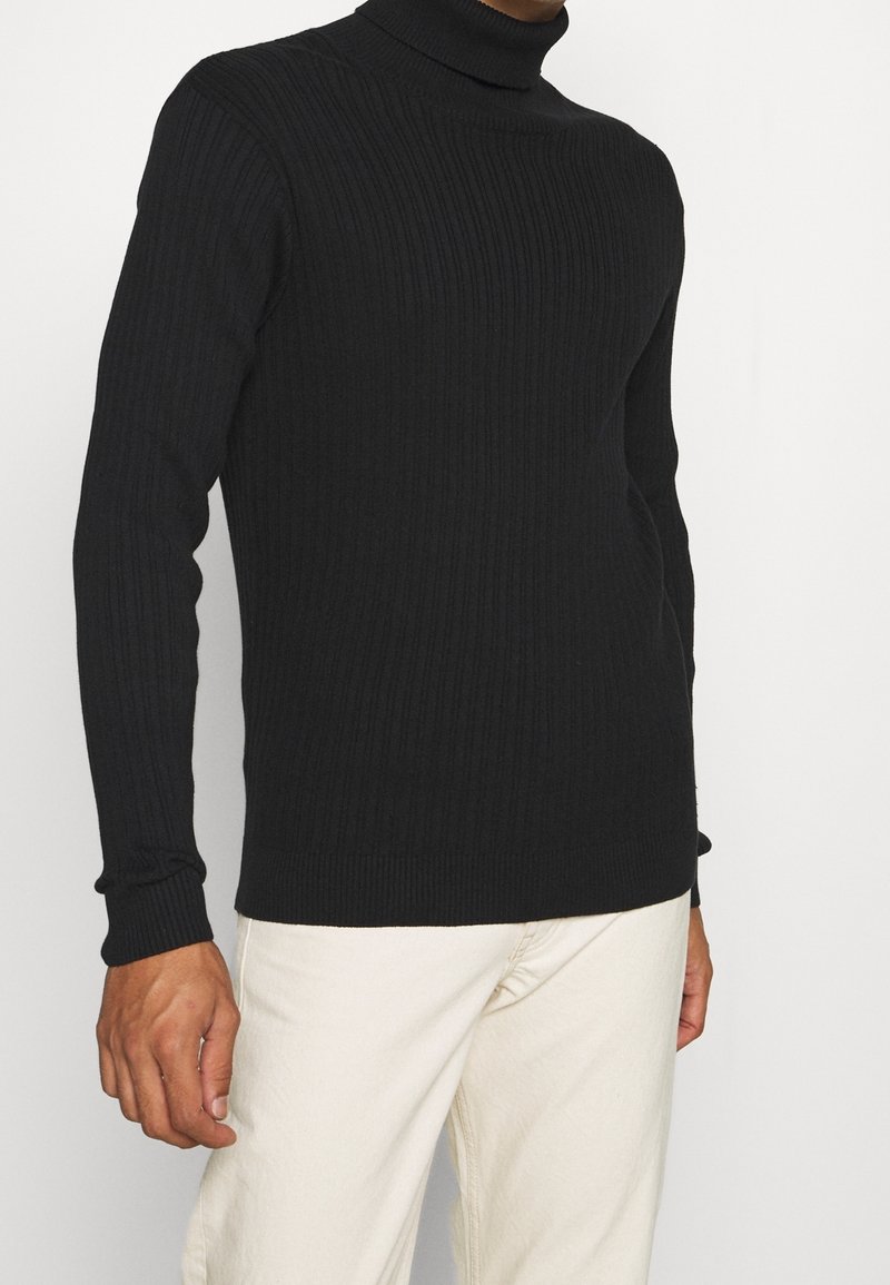 Homme portant un pull à col roulé côtelé noir et un pantalon blanc cassé, debout devant un fond clair uni.