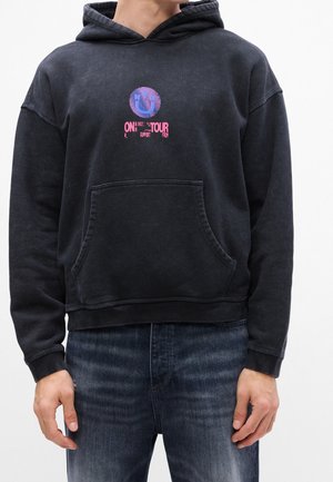 Persoon draagt een vervaagde zwarte hoodie met een paarse smiley en roze tekst 'ON TOUR' op de borst, gecombineerd met donkerblauwe spijkerbroek.