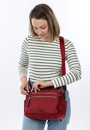 Woman in striped long-sleeve shirt and jeans unzipping a red crossbody bag against a white background.