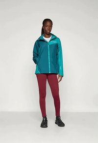 Vannbestandig teal jakke med full glidelås, hette og kontrastpanel på skuldrene. Kombinert med maroon leggings og svarte joggesko.