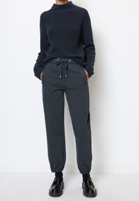 Maglione lavorato a maglia blu navy con collo alto, abbinato a pantaloni jogger grigi scuri a gamba affusolata con vita elastica e tasche laterali. Calzature nere.
