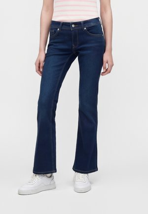 Personne portant un jean bootcut bleu foncé et des baskets blanches debout devant un fond uni.