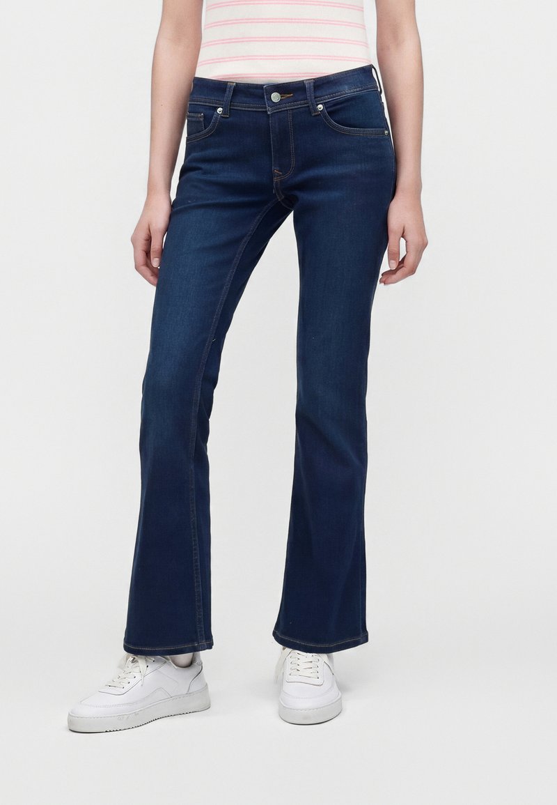 Άτομο που φοράει σκούρα μπλε τζιν bootcut και λευκά αθλητικά παπούτσια, στέκεται μπροστά σε απλό φόντο.