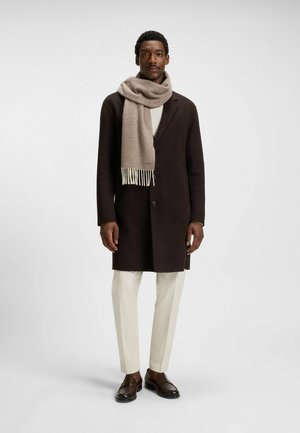 Cappotto marrone con una morbida sciarpa beige, maglione bianco e pantaloni chiari. Dettaglio della sciarpa con frange, scarpe lucidate e vestibilità su misura.