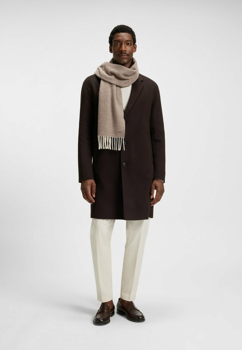 Bruine lange jas met een zachte beige sjaal, witte trui en pale broek. Sjaal met franjes, gepolijste schoenen en een passend model.