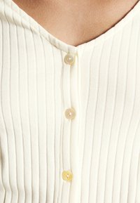 Krämfärgad ribbad topp med en V-ringning och tre ljusa knappar framtill, som framhäver ett texturerat tyg.