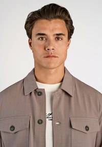 Brun knappad jacka med krage, två framfickor och svarta knappar, bärs över en vit skjorta med tryckt text. Slät, texturerad tyg.