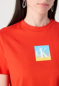 Rode katoen T-shirt met een blauw naar geel verloop rechthoekig logo met "cK" in het wit. Korte mouwen en ronde hals.