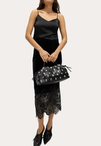 Zwarte leren handtas met zilveren studs, geplooid ontwerp en verstelbare trekkoorden. Model draagt een satijnen zwarte top en een rok met kantaccenten.
