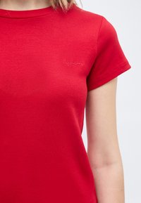 Κοντινή λήψη κόκκινης ριμπ μπλούζας με κοντά μανίκια και μικρό κεντημένο λογότυπο στο στήθος, που φοριέται από άτομο με ανοιχτόχρωμο δέρμα και ξανθά μαλλιά.